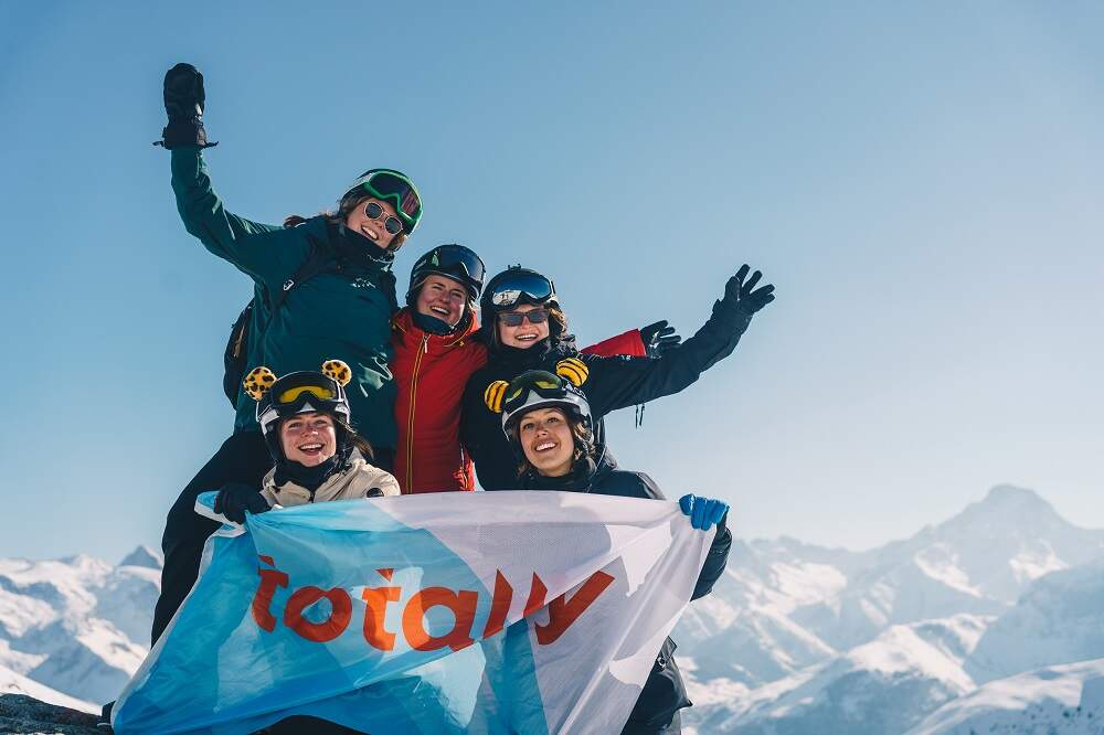 blije groep vrienden in skikleding met totally vlag in hun handen boven op een berg in een zonnig skigebied