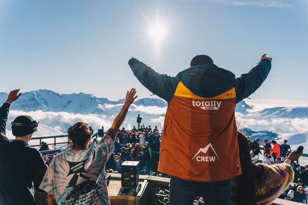 Totally crew member die in dj boot staat naast dj's met armen in de lucht tegenover een groep feestende mensen in skigebied