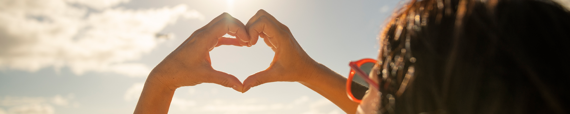 Vrouw of meisje dat een hartje maakt met haar handen richting de zon met een zonnebril op en bruin haar