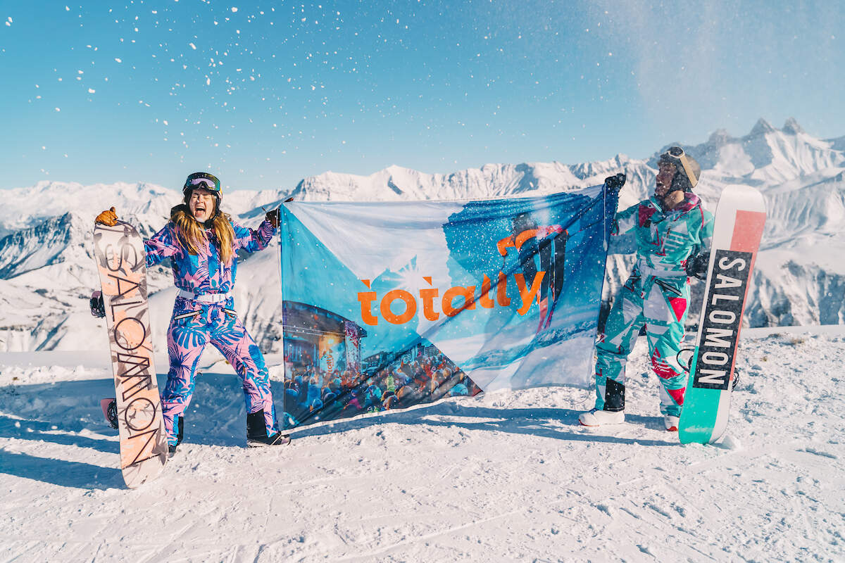 Man en vrouw die samen een vlag van Totally snow vasthouden op een besneeuwde bergtop