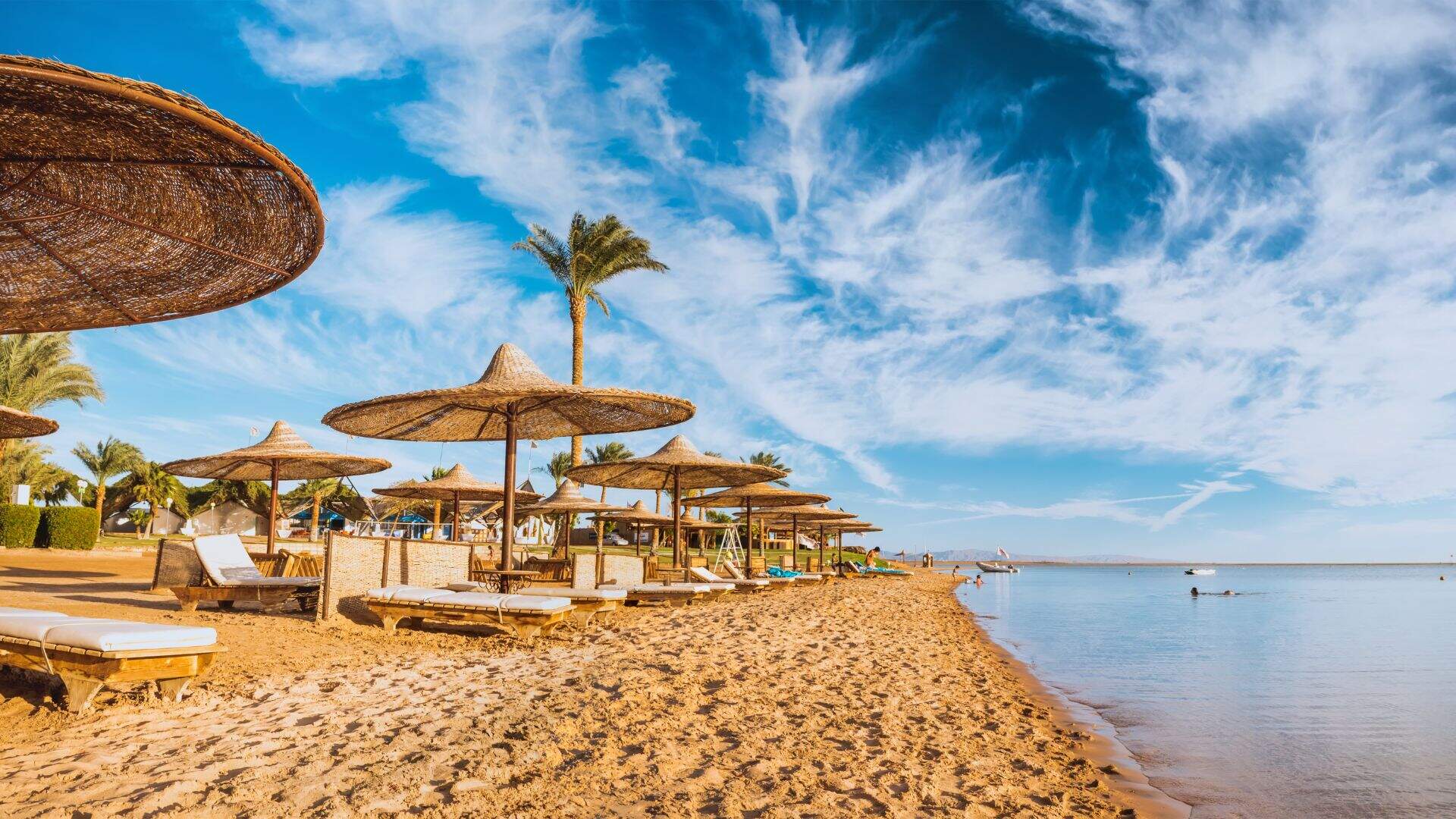 zandstrand aan kalme zee met bewolkte lucht, rieten parasollen en ligbedjes met kussens in Egypte