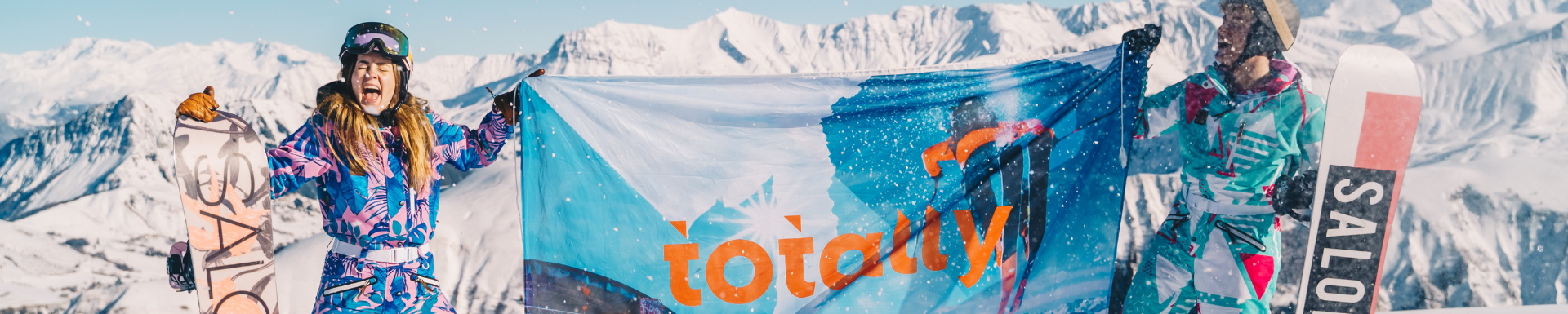 man en vrouw die lachend een totally snow vlag hoog in de lucht houden op besneeuwde bergtop