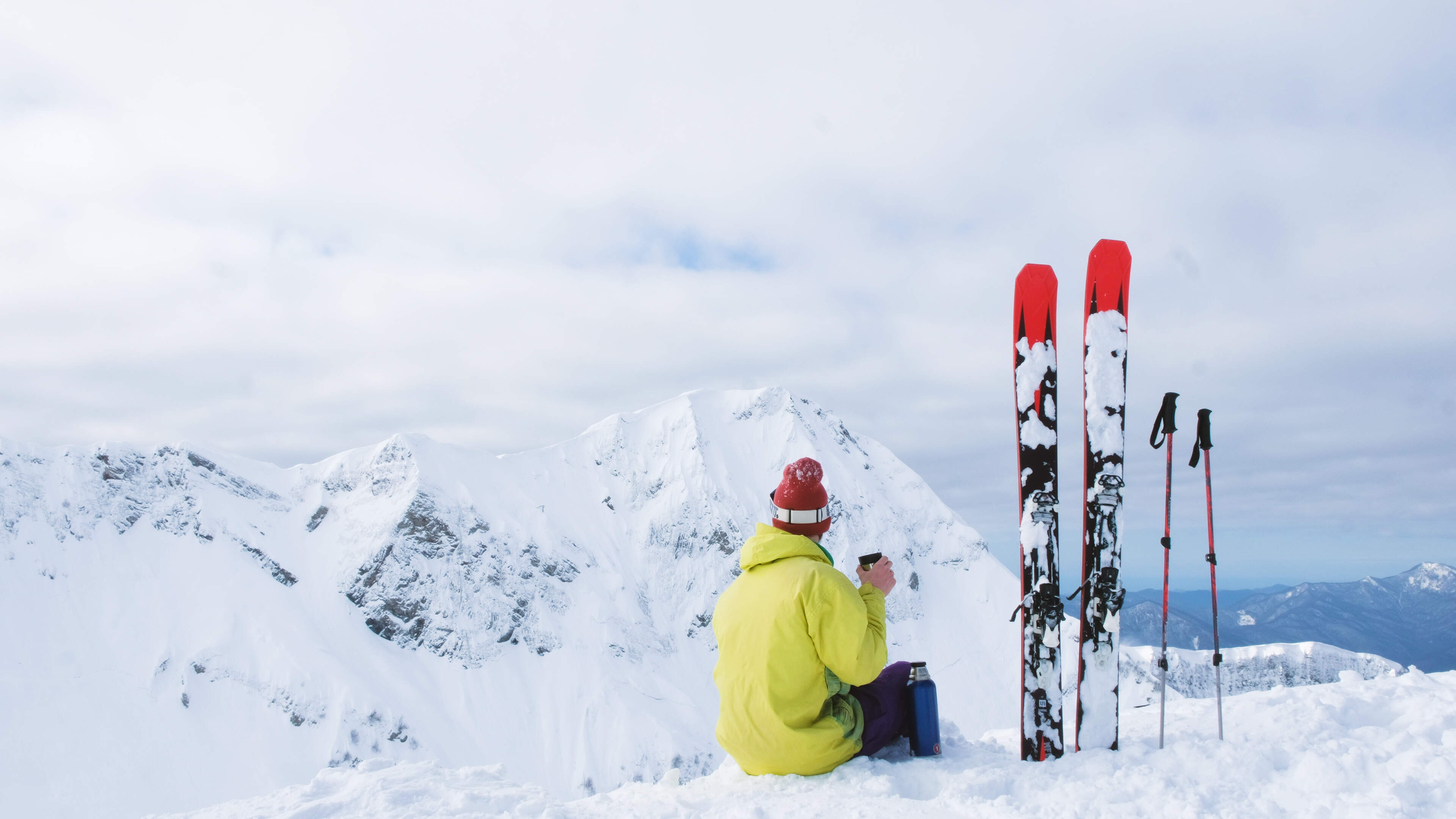 skier met gele jas zit op top van piste met een beker in zijn hand en kijkt uit over de besneeuwde bergtoppen
