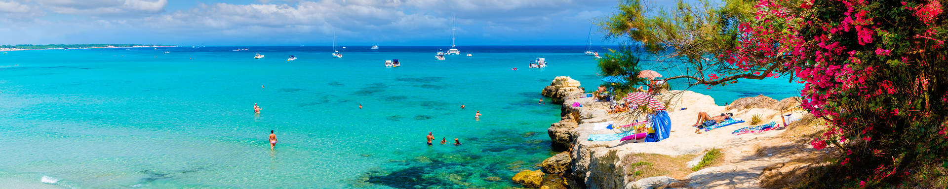 Uitzicht over azuurblauwe zee met boten, zwemmende mensen en een strakblauwe lucht. 